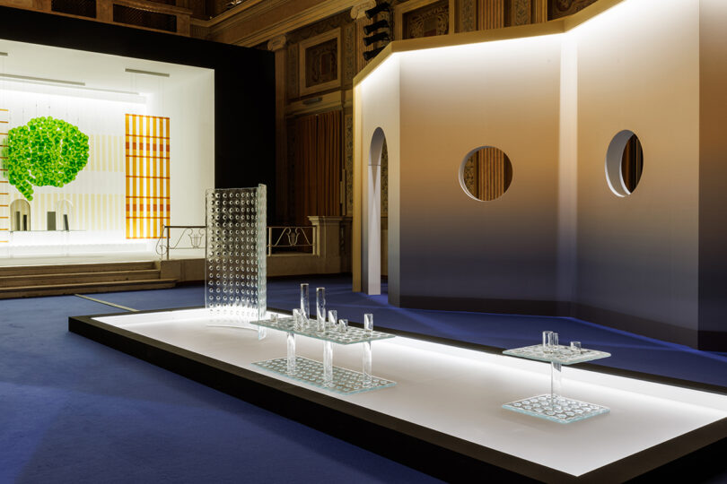Contemporary exhibition space with geometric sculptures, glass objects on display tables, and illuminated architectural elements against ornate walls.