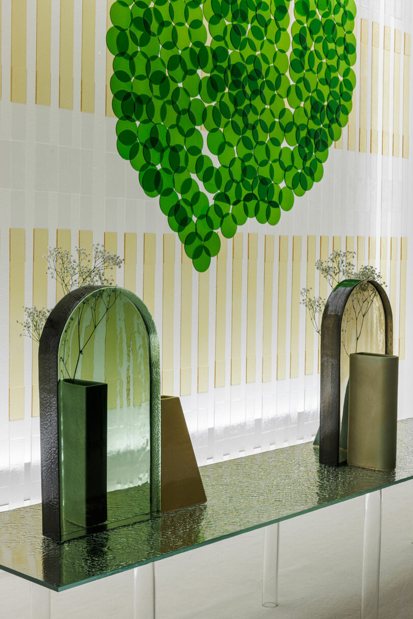 A glass table with minimalist vases and mirrors from POETICA Wonderglass sits before a wall adorned with a green circular leaf design and vertical beige stripes.