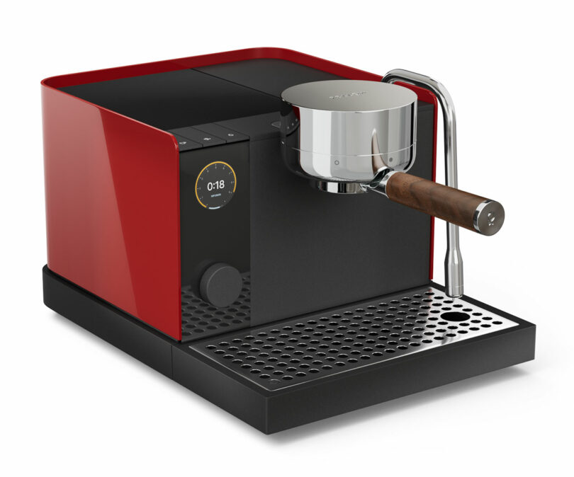 A red espresso machine with Fellow coffee-making tools and a milk frothing pitcher sits on a wooden counter against a textured wall, sunlight streaming through the window.