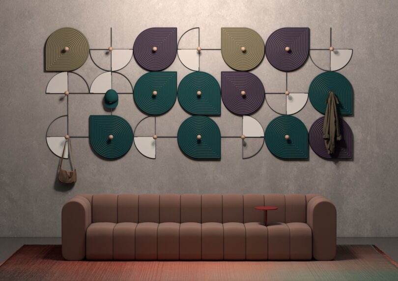 Modern living room with a mauve sofa and a colorful geometric wall art installation. A small red tray is attached to the sofa arm. A bag and a coat hang on the wall art