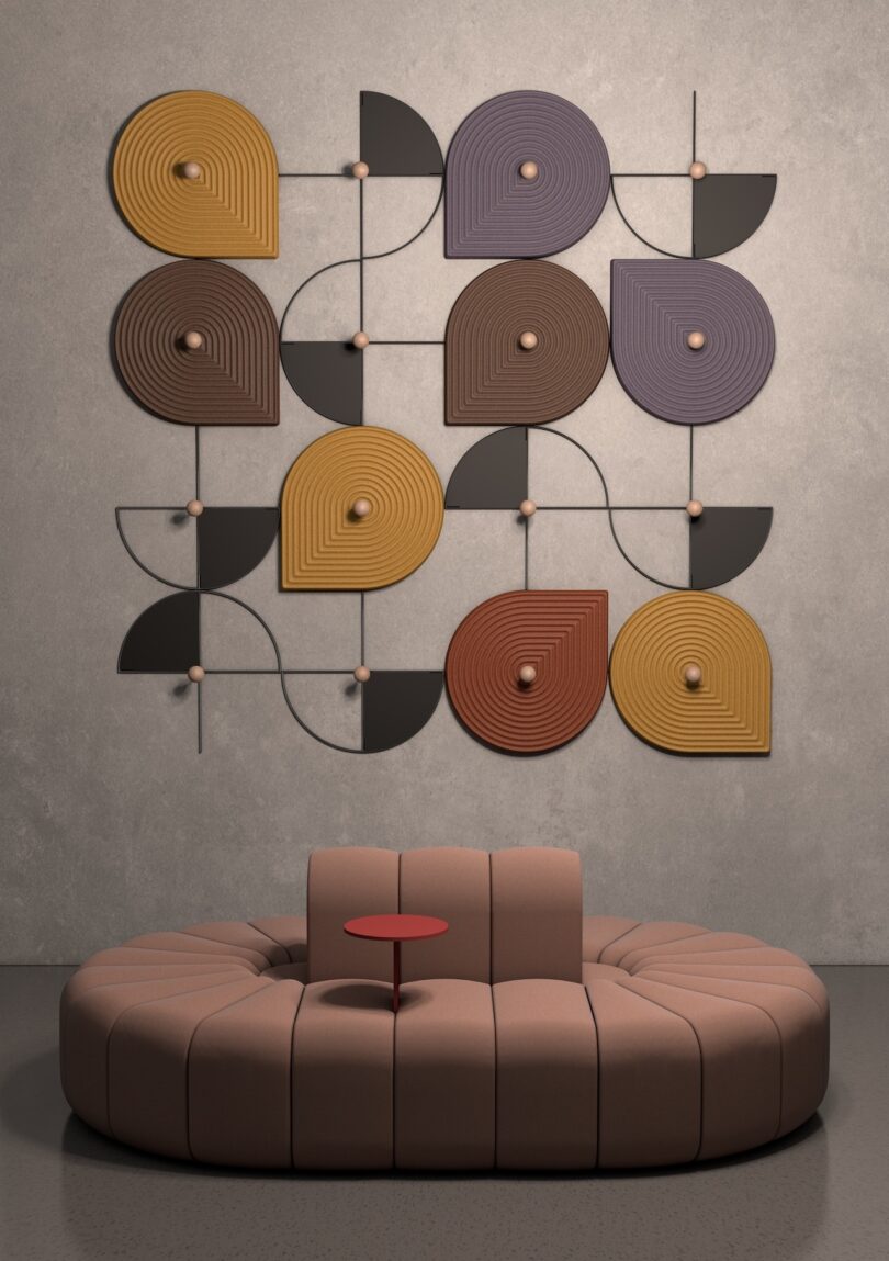 A circular beige sofa with a small red table in the center is in front of a wall art piece featuring geometric shapes in earthy tones