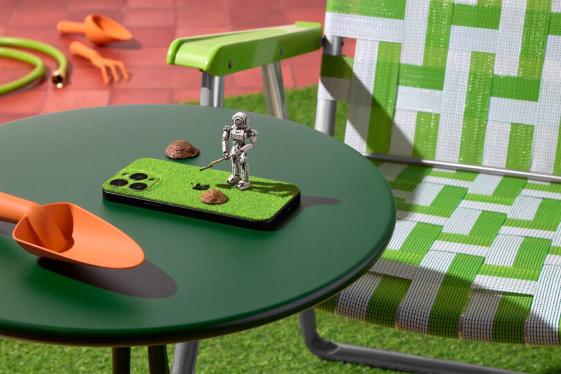 A green round table with an orange trowel and a phone decorated with fake grass, rocks, and a small robot figure, next to a green-and-white lawn chair.