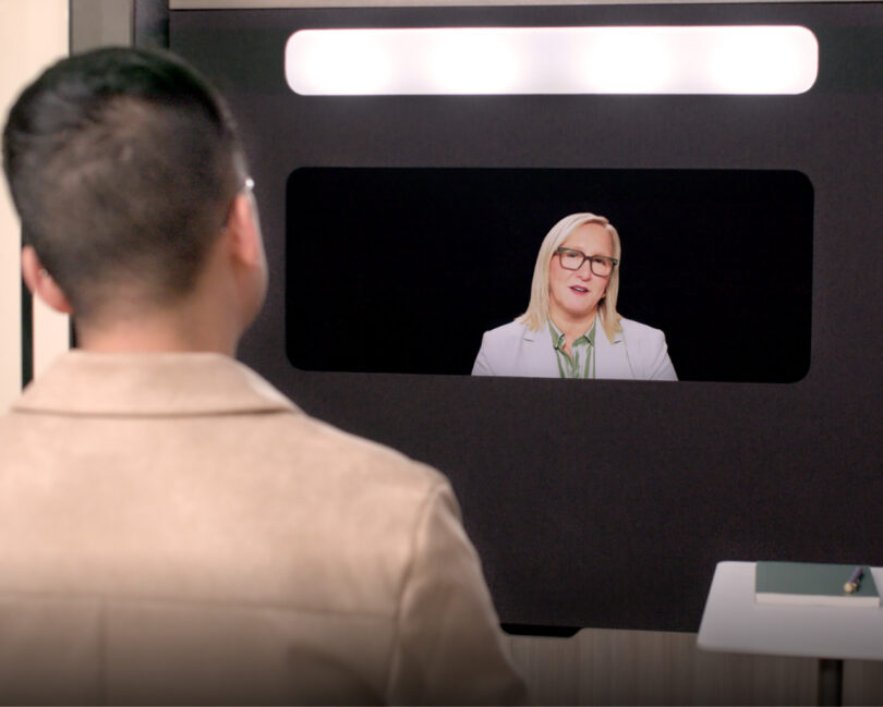 Blond woman with glasses in professional attire on the Ocular View screen speaking to a man with short hair as seen from behind,