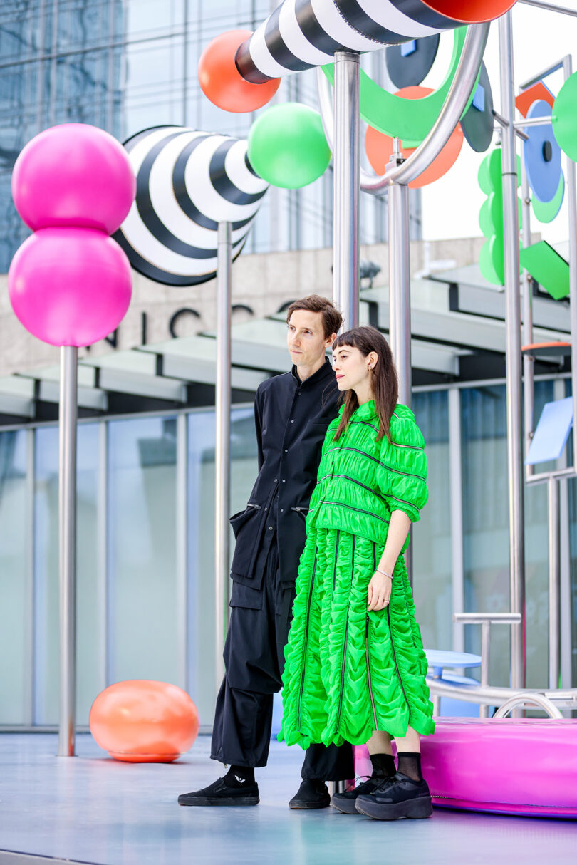 Two people stand side by side in front of colorful, abstract outdoor sculptures; one wears a black outfit, the other a bright green dress.