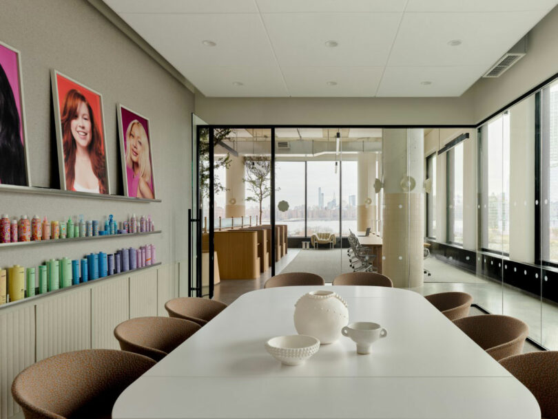 A modern meeting room with a white table, brown chairs, white ceramic vases, colorful bottles on a shelf, and portrait photos on the wall; glass doors open to another office area.