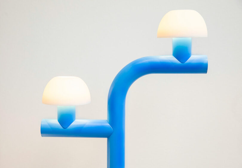 A modern lamp with a blue, Y-shaped base and two white, dome-shaped light shades set against a plain background, inspired by Nader Gammas’ contemporary design sensibility.