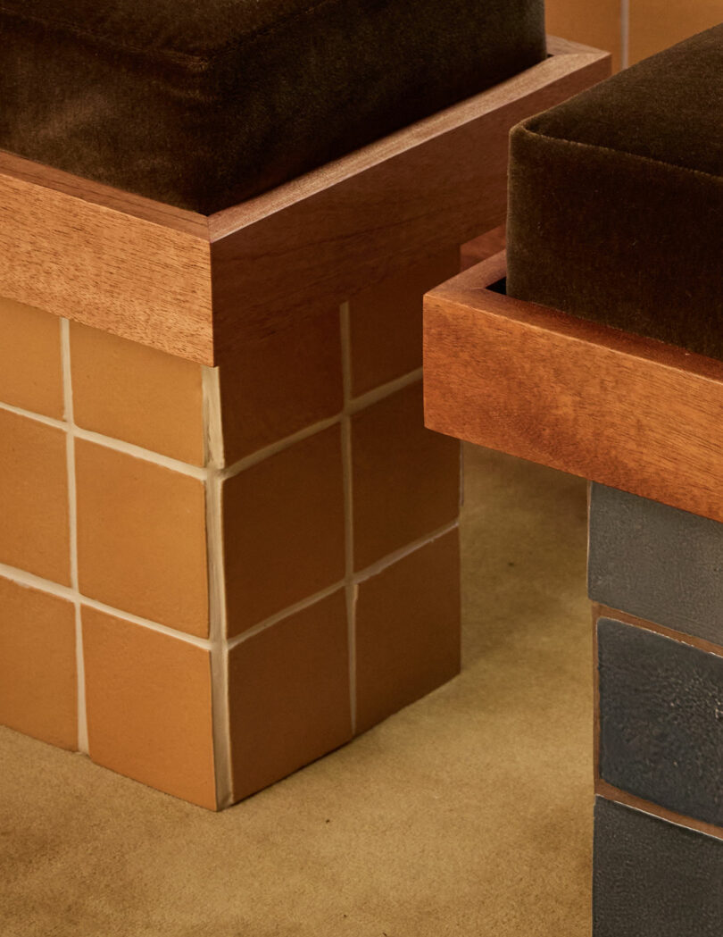 Two square wooden stools with velvet cushions; one base is tiled in tan squares, the other in dark blue tiles—an eye-catching pair from Ah Um, placed on a beige carpet.