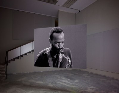 A video projection of a Black man playing a saxophone shown on a hanging screen in an empty gallery.