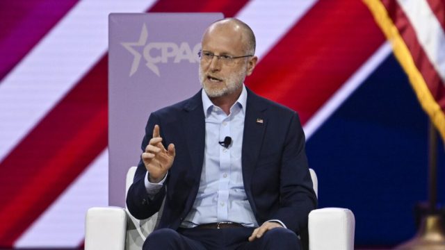 FCC-Chair-Brendan-Carr-is-letting-ISPs-merge—as-long-as.jpg Federal Communications Commission member Brendan Carr sits on a stage and speaks while gesturing with his hand. Behind him is the CPAC logo for the Conservative Political Action Conference.