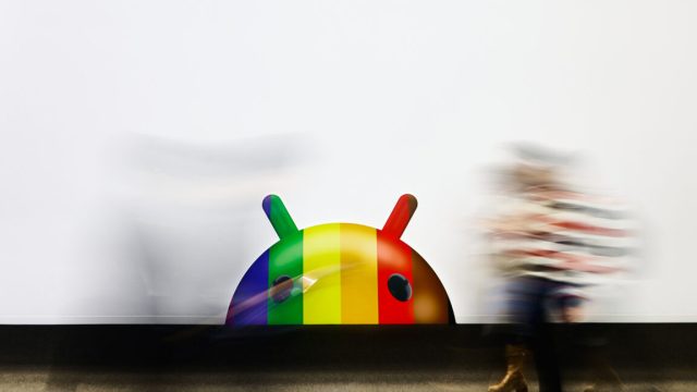 Google-wanted-that-Nextcloud-decries-Android-permissions-as-gatekeeping.jpg Android robot mascot, its head in rainbow stripes, peeking above a white background horizon, with people blurred in motion moving past.