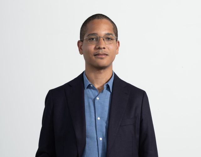 A young man with glasses and short hair in a blue button up shit and dark blue blazer.