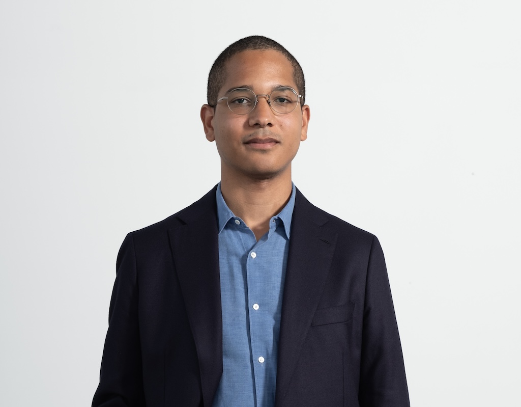 A young man with glasses and short hair in a blue button up shit and dark blue blazer.