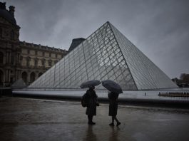 Two people shelter under an umbrella during a rainy day next to a glass pyramid structure.