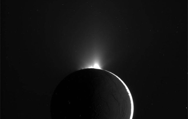Testing-a-robot-that-could-drill-into-Europa-and-Enceladus.jpeg A greyscale image of a crescent view of a moon, with a bright spike coming out of its top.