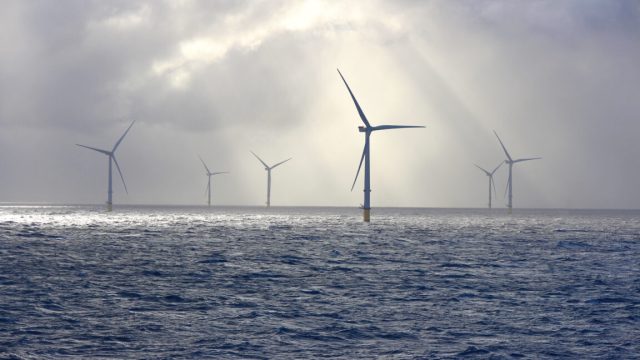 Trump-admin-lifts-hold-on-offshore-wind-farm-doesnt-explain.jpg A series of tall wind turbines in the ocean, illuminated by beams of sunlight filtering through baps of heavy clouds.