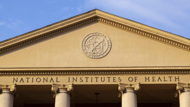 Trumps-NIH-ignored-court-order-cut-research-grants-anyway.jpg A view of the exterior of a building with a triangular top supported by pillars. Beneath a circular logo are the words "national Institutes of Health"