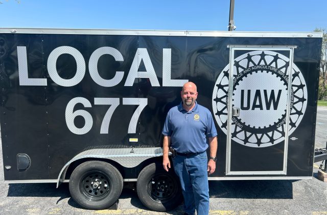 Trumps-tariffs-are-already-destroying-jobs.jpeg Hand, a bald white man with a neat grey goatee, wears a blue UAW polo and stands in front of a trailer that has the UAW seal and the words “Local 677” on it.