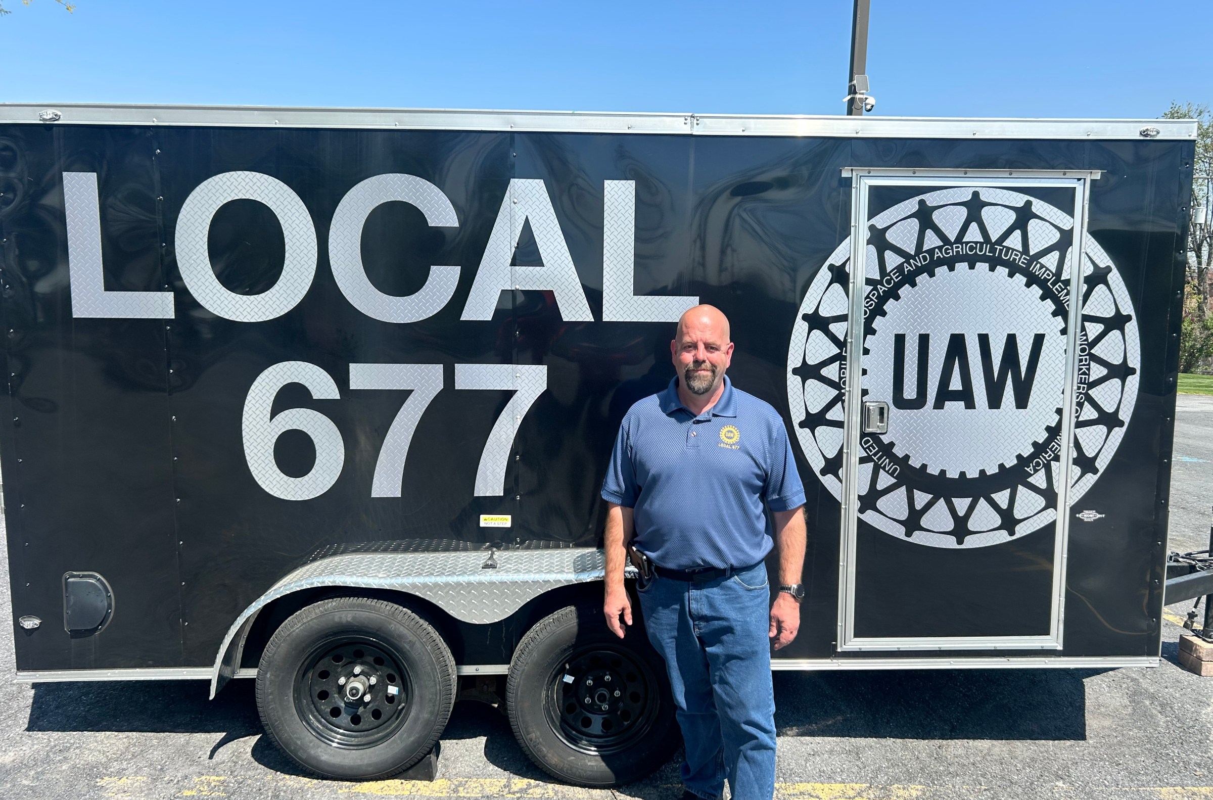 Trumps-tariffs-are-already-destroying-jobs.jpeg Hand, a bald white man with a neat grey goatee, wears a blue UAW polo and stands in front of a trailer that has the UAW seal and the words “Local 677” on it.