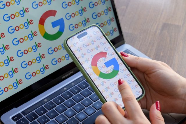 US-DoJ-wants-Google-to-sell-two-of-its-ad.jpg A woman looks at a mobile phone displaying the logo of Google in front of a laptop screen displaying the logo of Google.
