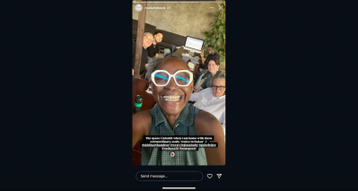 A Black woman taking a selfie with five other people seated at a table behind her.