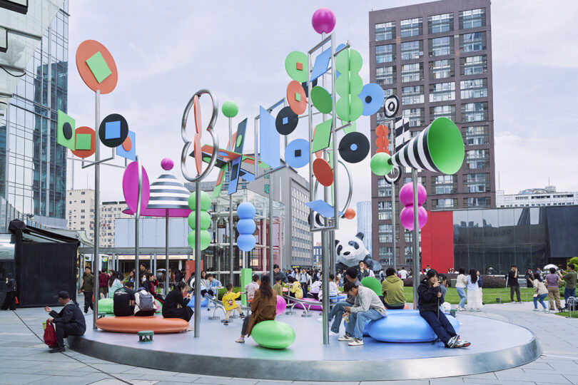 People gather and sit around a colorful, modern outdoor art installation with geometric shapes and tall buildings in the background.