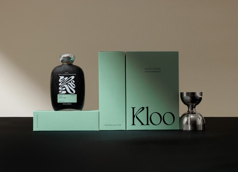 A bottle of Kloo craft coffee concentrate with minimalist green packaging and a silver cup arranged on a black surface against a neutral background.