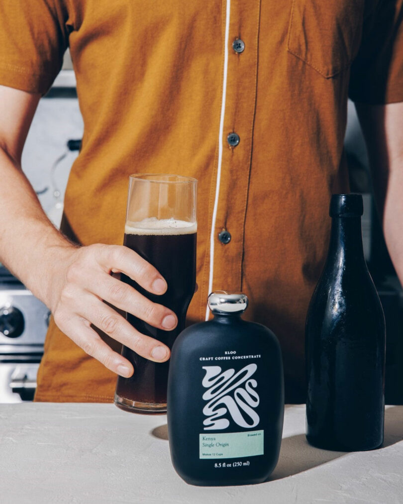 A person in a brown shirt holds a tall glass of dark beverage next to a black bottle and a black bottle labeled 