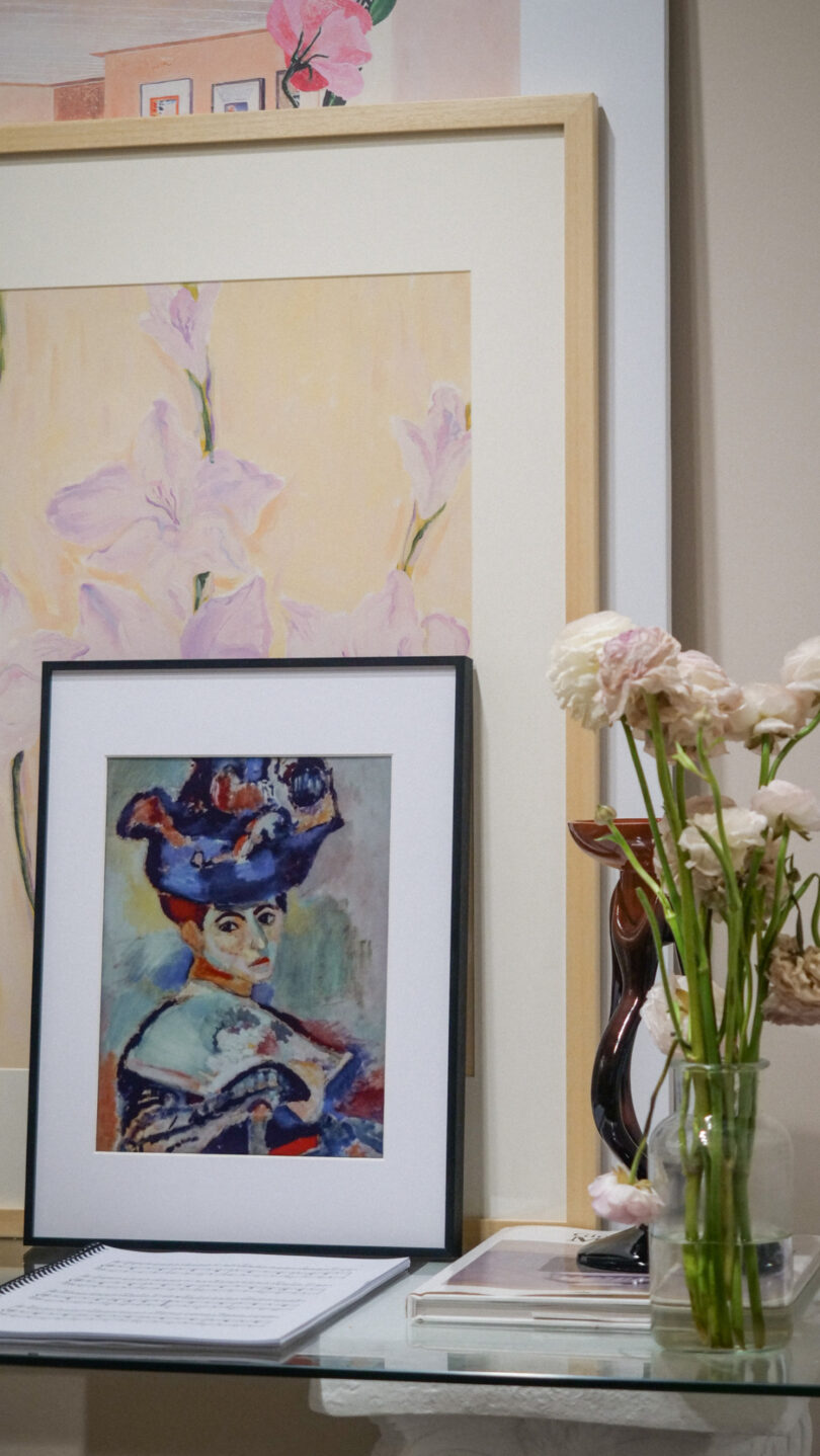 Framed paintings and a vase of light pink flowers are arranged on a table next to sheet music and books.