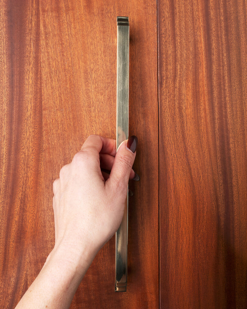 A hand gripping a vertical brass handle on a wooden cabinet door
