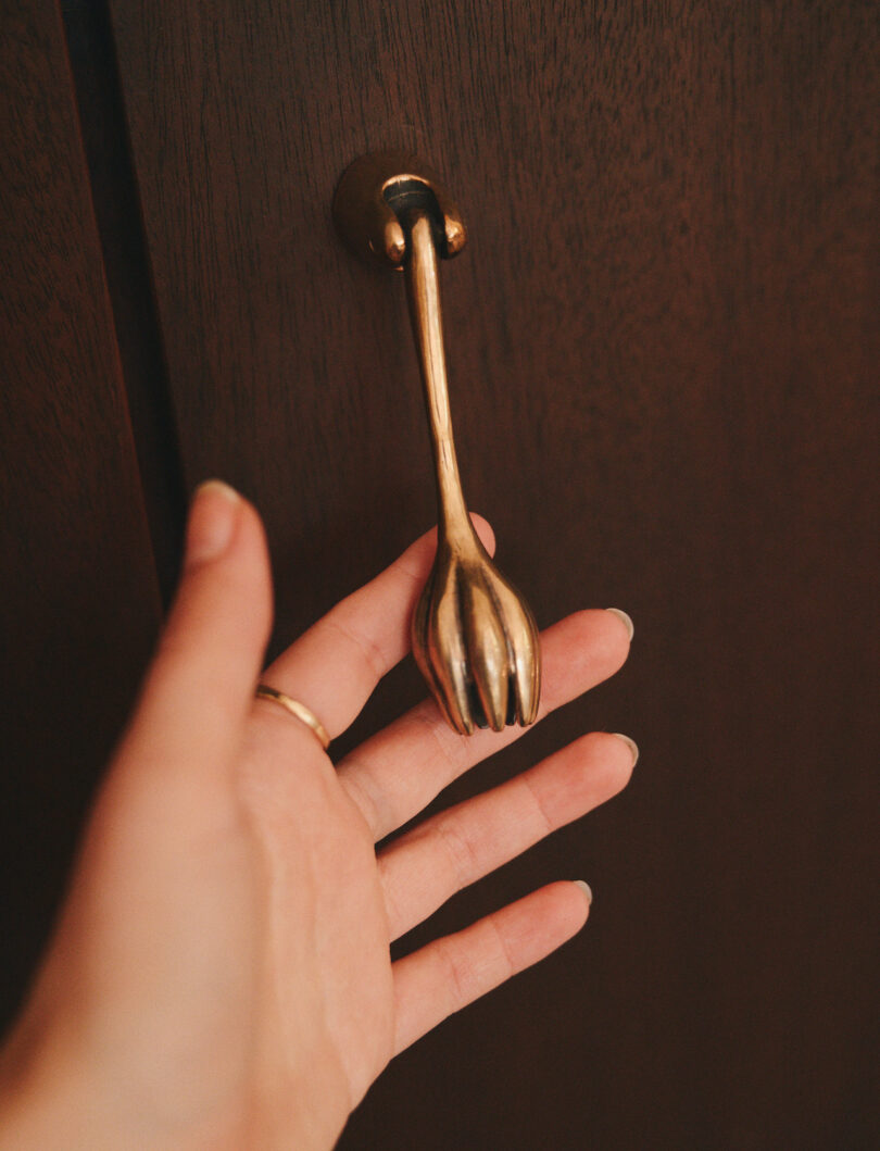 A hand reaches for a cabinet door handle shaped like a small brass fork against a dark wooden surface