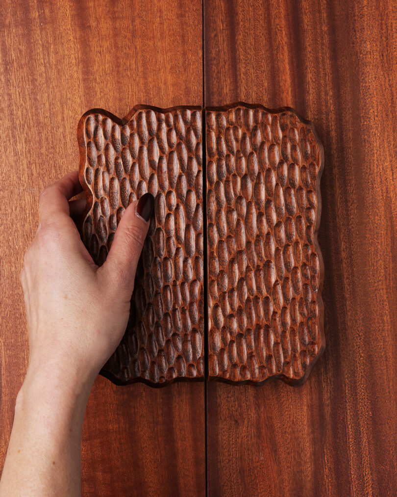 A hand with dark nail polish grasps a textured wooden handle on a wooden cabinet with a matching carved design