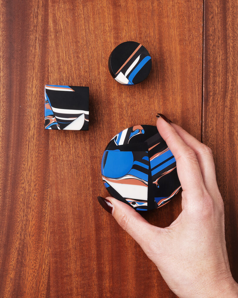 A hand holds a round, abstract-patterned object with blue, black, white, and brown swirls; two matching smaller objects rest on a wooden surface