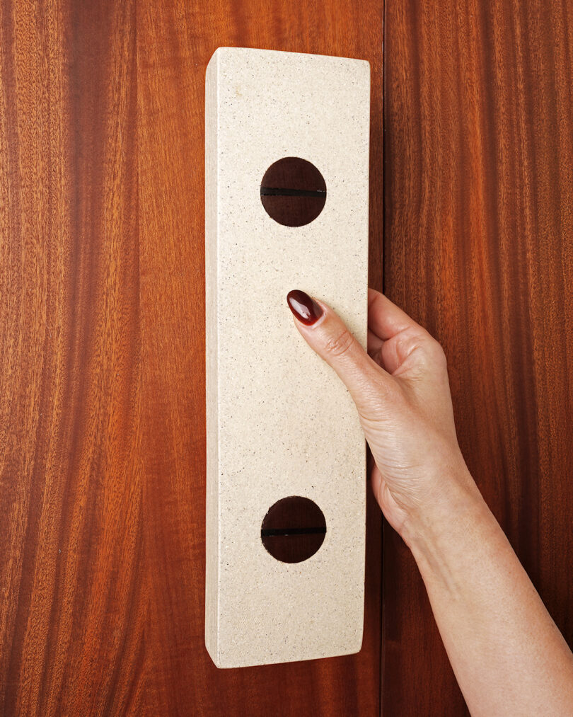 A hand with red nail polish holds a rectangular beige object with two circular holes against a wooden surface