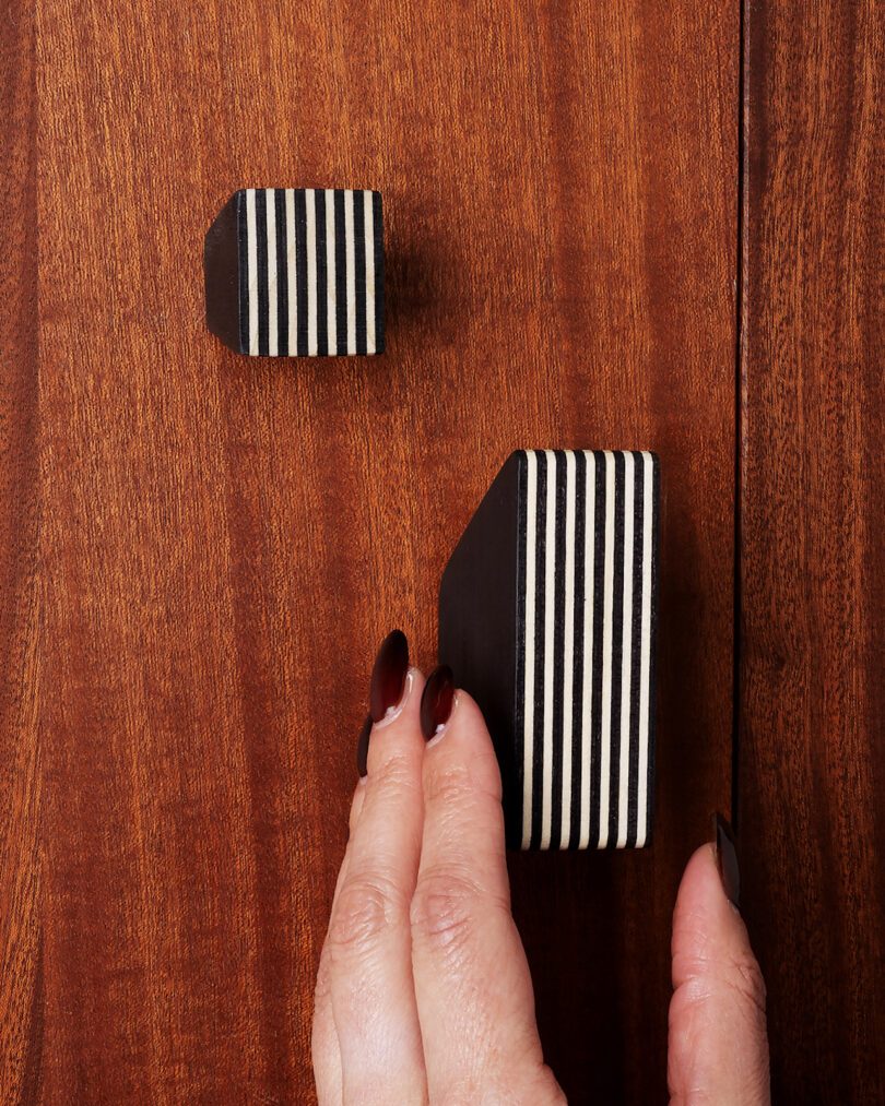 A hand with painted nails grasps a striped black and white cabinet knob on a wooden surface, with a smaller matching knob visible above