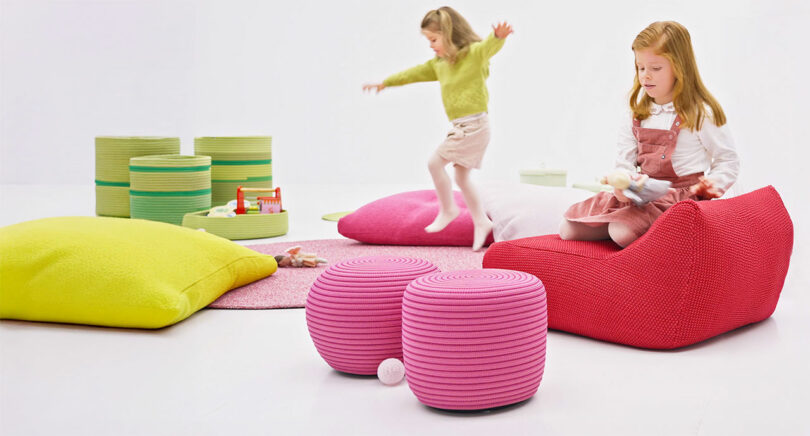 Two young girls play in a brightly colored room featuring pink and green cushioned furniture from the Paola Lenti Kids Collection, with toys scattered on the floor.