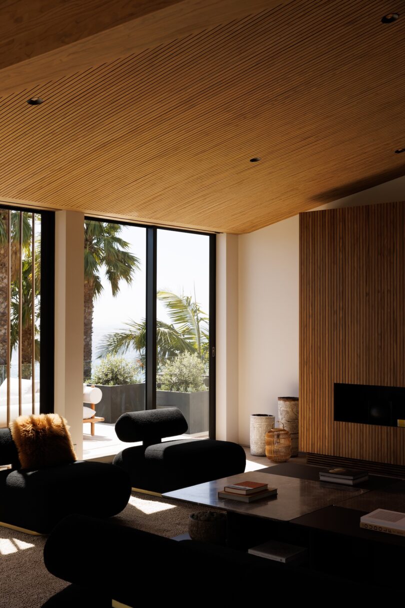 Modern living room with black sofas, wooden ceiling and wall panels, large windows, and a view of palm trees and outdoor seating. Natural light fills the space.