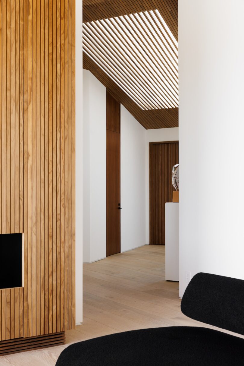 Modern interior with vertical wood paneling, a slatted wood ceiling feature, white walls, light wood floor, black chair, and a closed wooden door in the hallway.