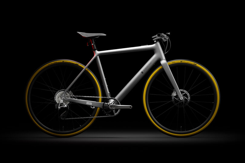 A sleek, modern bicycle with a silver frame, yellow-rimmed tires, and disk brakes, displayed against a black background.