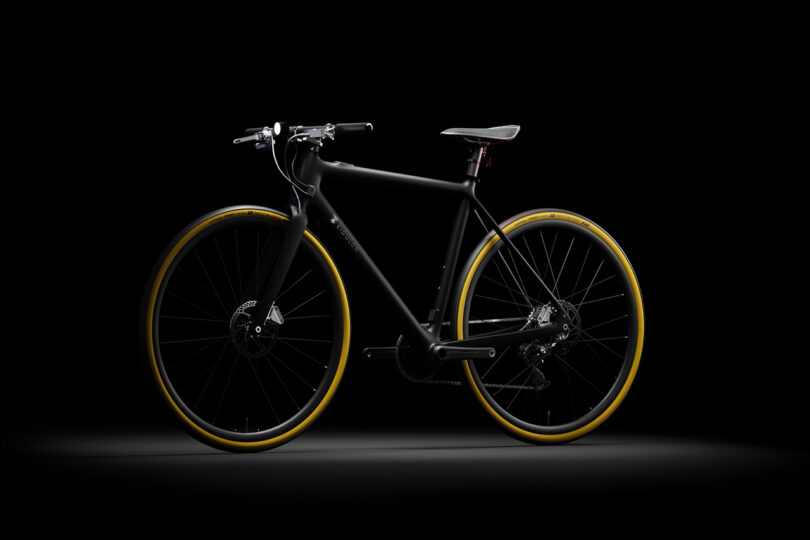 A sleek black bicycle with yellow tires is displayed against a dark background, highlighting its streamlined design and modern features.