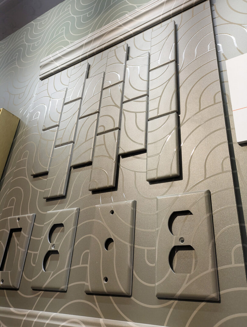 A display of various metallic wall plates for light switches and electrical outlets, arranged on a patterned wall, showcases the NeoCon 2025 Top 10 design innovations.