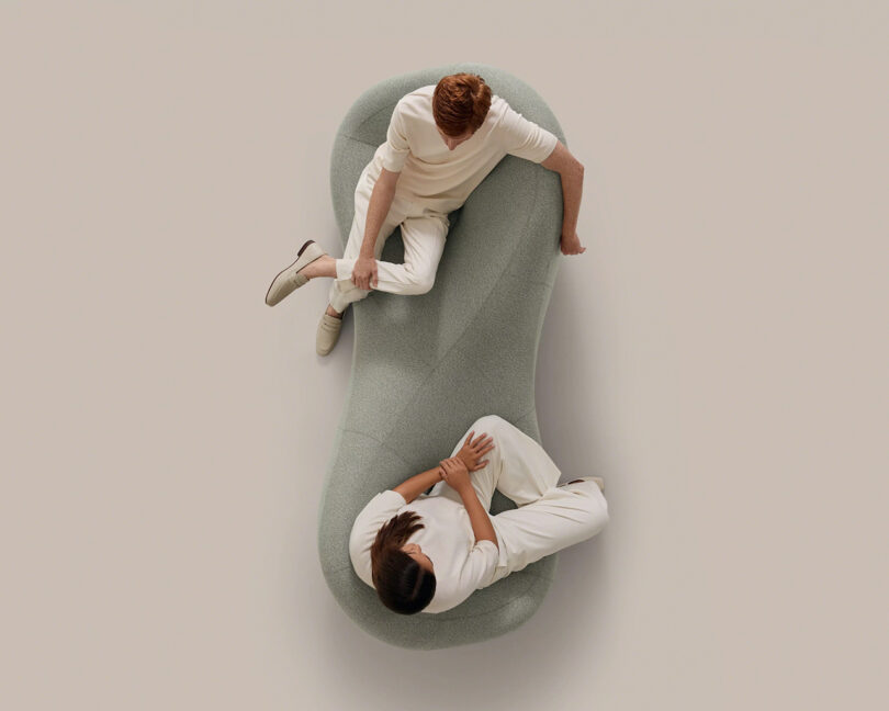 Two people dressed in white sit on a bean-shaped light gray sofa, facing each other and talking, viewed from above against a plain background.