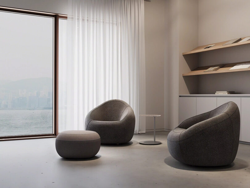 Minimalist room with two gray armchairs, a round ottoman, a small side table, large window with sheer curtains, wall shelves, and a view of water and distant buildings.