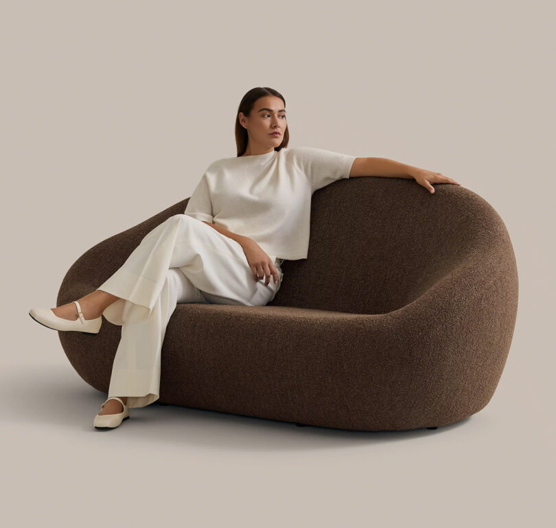 A woman in white clothing sits on a modern, brown, curved sofa against a plain beige background.