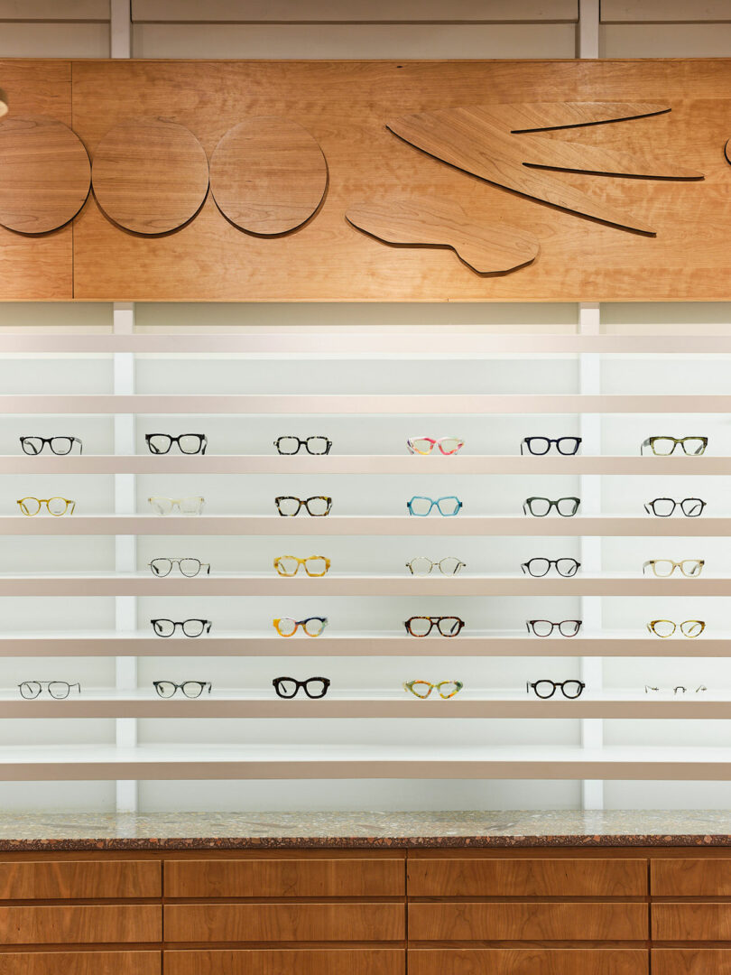 A display of eyeglasses on horizontal shelves with a wooden backdrop and abstract wooden art pieces above.