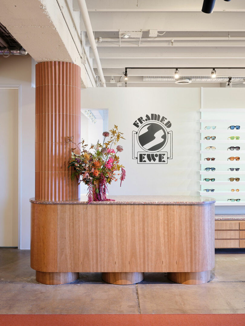 A modern eyewear store interior with a curved wooden counter, a floral arrangement, and a wall display of various glasses. The store logo, 