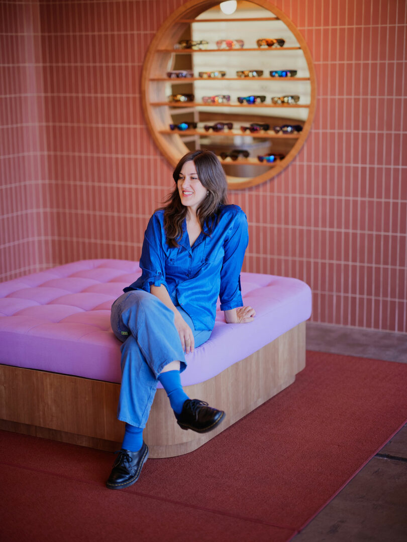 A person in a blue outfit sits on a purple cushioned bench in a store with a circular shelf displaying glasses in the background.