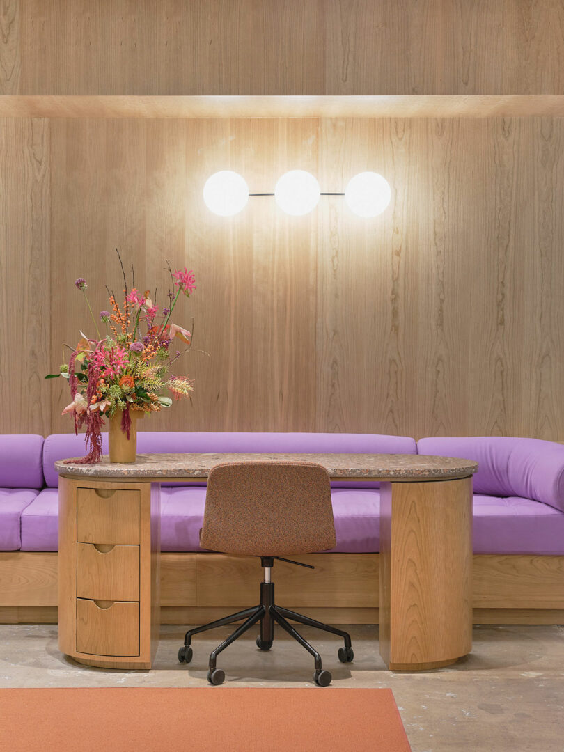 A modern office setup with a wooden desk, an upholstered office chair, a vase of flowers, and a purple sofa. Three round lights are mounted on the wooden wall behind.