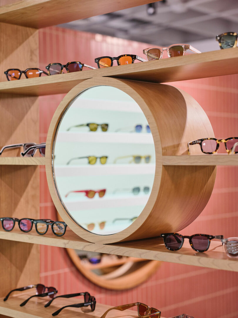 A wooden shelf display with various eyeglasses surrounds a circular mirror, reflecting more eyewear in a store setting.