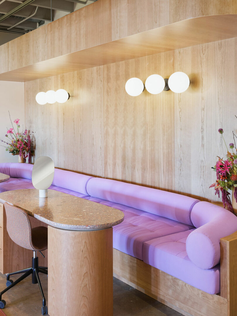 Modern waiting area with a purple cushioned bench, round wall lights, a wooden desk with a mirror, and decorative flowers.