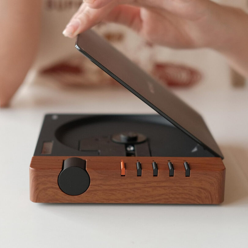 A person opening the lid of a Syitren R300 compact CD player with a wood-like finish. The player features a large round knob and several switches, offering both style and functionality.